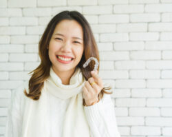 Brunette Asian woman in a white sweater smiles as she holds her Invisalign trays against a white brick wall