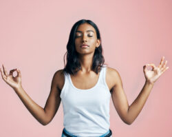 A Brown woman in a white tank top closes her eyes and holds her hands in a zen pose as she tries to relax